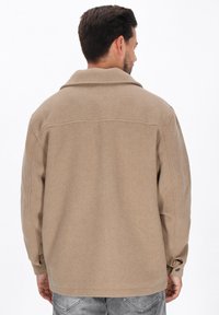 Beige wolmixjas met een puntige kraag, voorzien van een rechte snit, manchetknoepen en een soepele textuur, weergegeven vanuit de achterkant.