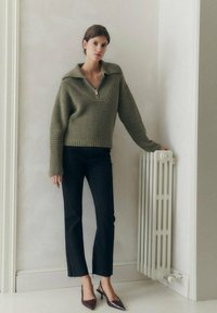 Pullover verde lavorato a maglia con colletto ampio, zip frontale a mezza lunghezza e texture a coste, abbinato a pantaloni neri cropped e tacchi a punta.