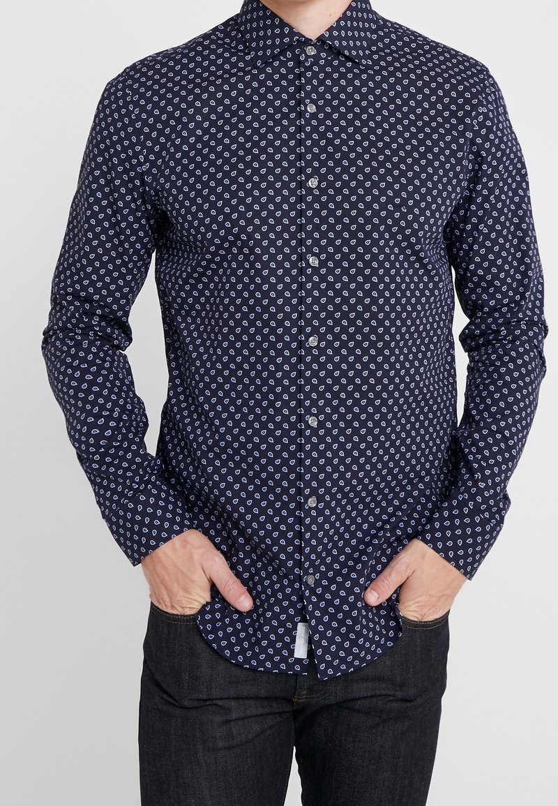 Camicia azzurra navy con un piccolo motivo di cuori bianche, maniche lunghe e colletto a punta, abbinata a jeans scuri. Tessuto dalla texture morbida.