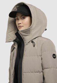 Beige pufferjack met een gewatteerde capuchon, contrasterende zwarte rits en een Khujo logo patch op de mouw. Gladde textuur, getailleerde pasvorm.