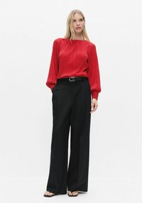 Rød plisseret bluse med lange ærmer og rund hals, kombineret med sort, højtaljede bukser med vide ben og flade sandaler.