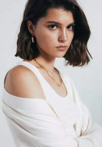 Bílý žebrovaný top bez ramínka s svetr na jedno rameno, doplněný vrstveným zlatým náhrdelníkem s různými texturovanými články.