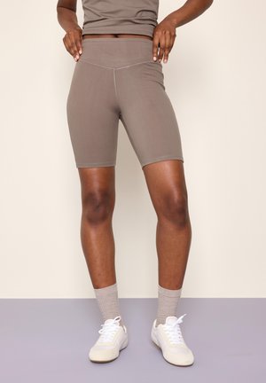 Person iført taupe bikershorts med høj talje, beige sokker, hvide sneakers og matchende cropped tanktop mod en neutral baggrund.