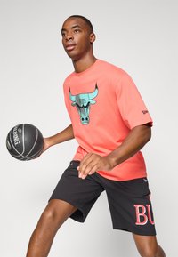 Korálové bavlnené tričko s grafiku tehlového býka, doplnené čiernymi šortkami s nápisom "BULL" v červenej farbe. Drží čierny basketbal.