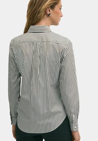 Pruhovaná košile v černobílé barvě, dlouhé rukávy, s klasickým límcem a detailním zapínáním na manžetách, se zakřiveným spodním lemem.