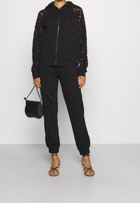 Žena v černé mikině na zip s krajkovými rukávy, k tomu ladící teplákové kalhoty s krajkovými panely, drží černou kabelku a má na nohou černé sandály se pásky.