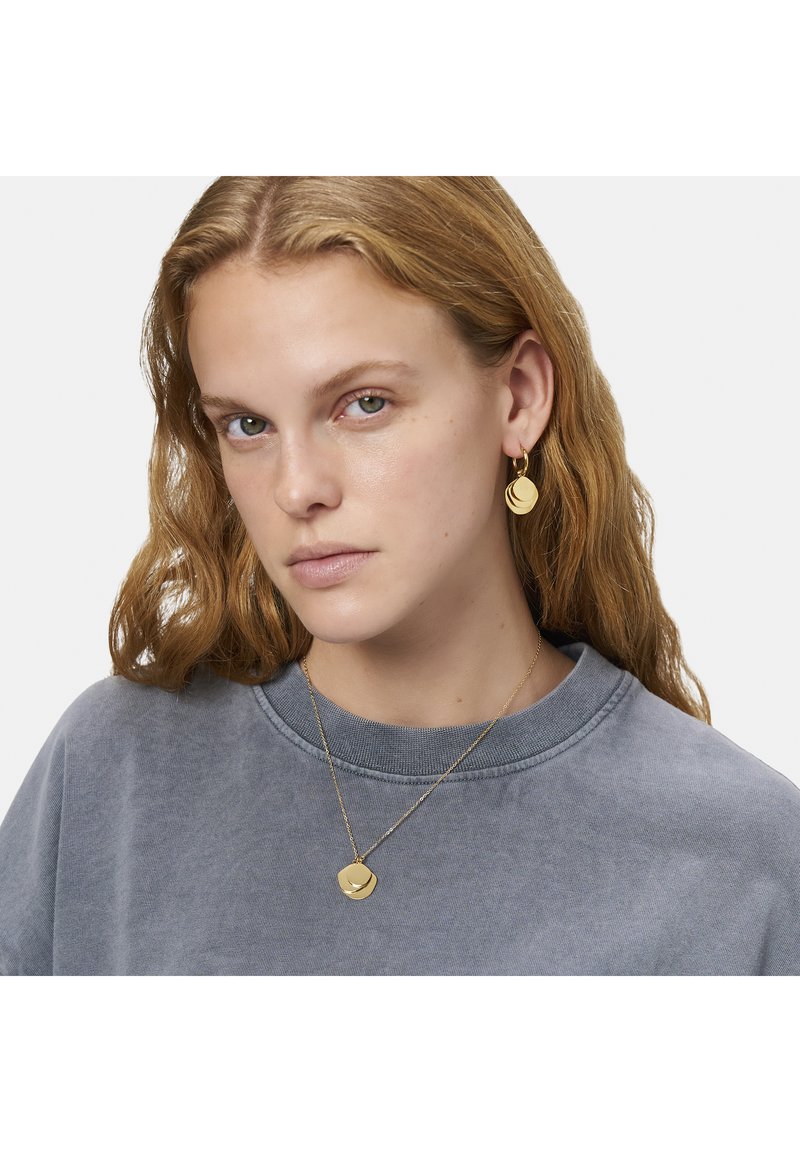 Gouden oorbellen met gelaagde ronde schijven en een bijpassende pendant ketting, gedragen over een grijze fleecetrui met een ronde hals.