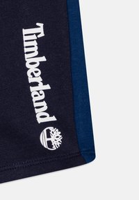 Mörkblå tygshorts med kontrasterande ljusblå sidpaneler, med "Timberland"-logotyp och träd-emblem i vit tryck.