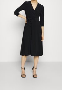 Černé šaty s obálkovým prálem a tříčtvrtečními rukávy, vypasovaný pás s mašlí a splývavou sukní. Lesklé černé lodičky s pásky kolem kotníků dokončují vzhled.