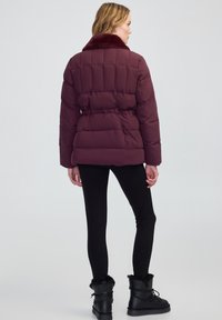 Donna che indossa un giubbotto trapuntato bordeaux con colletto di pelliccia, leggings neri e stivaletti neri foderati in pelliccia, in piedi voltata di spalle.