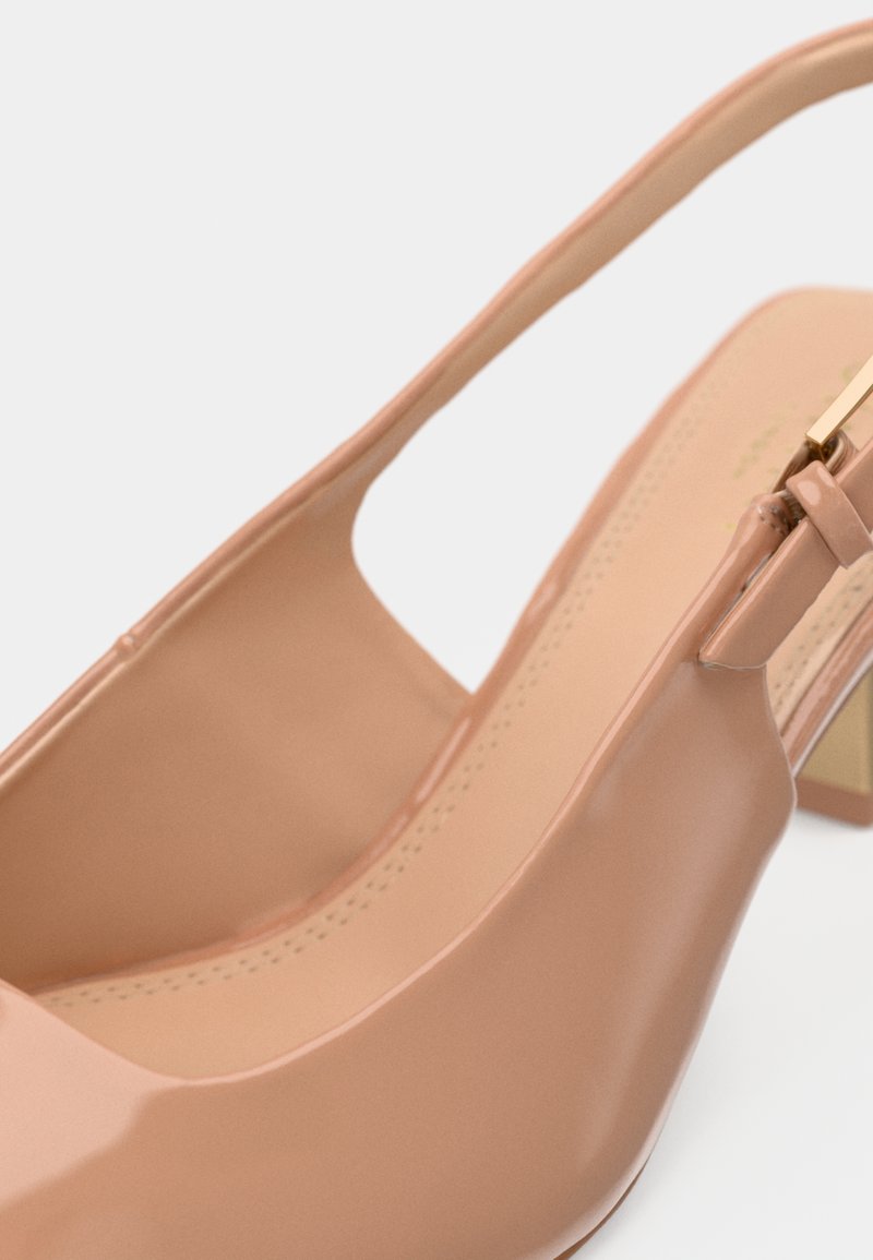 Escarpin en cuir verni beige avec un talon carré et une bride à boucle dorée, présenté en gros plan sur un fond blanc.