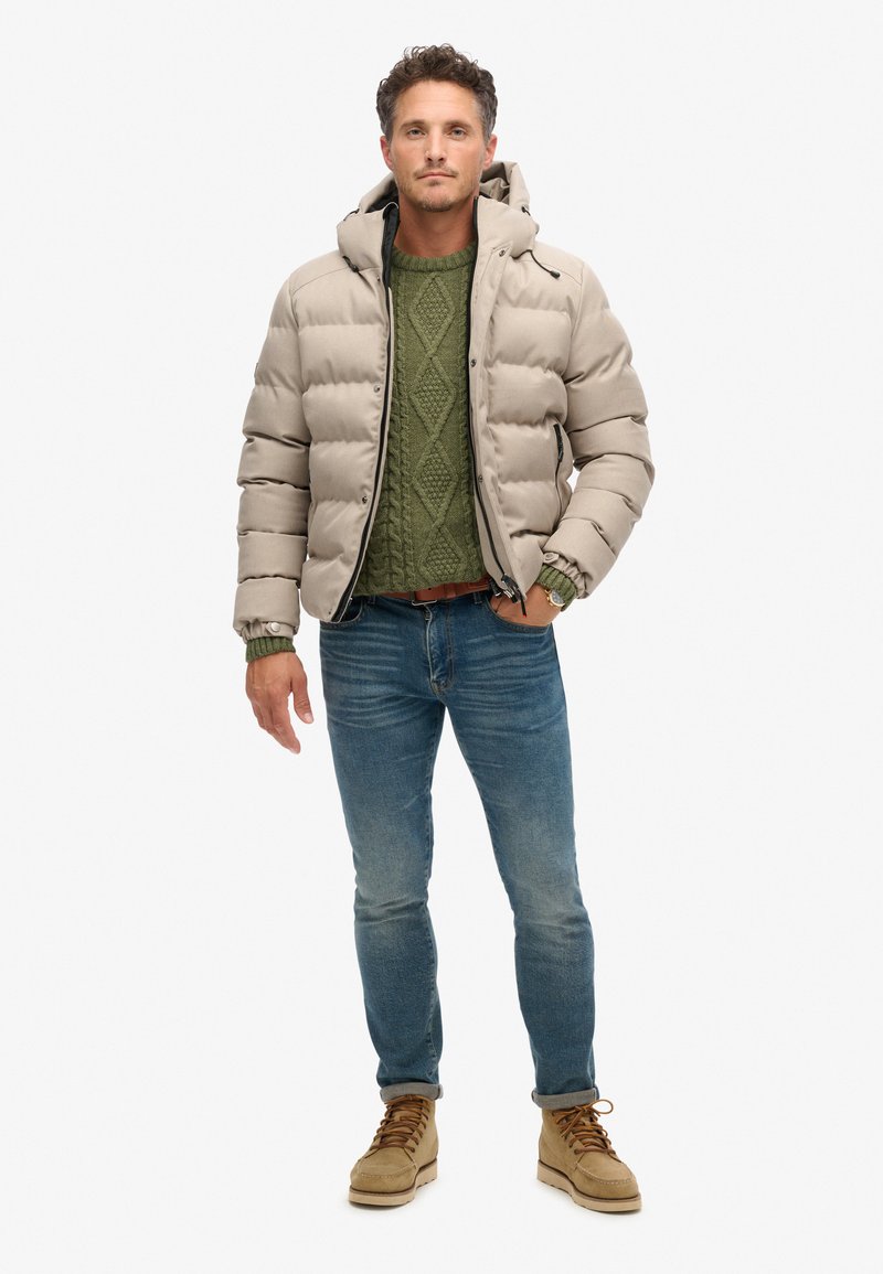 Beige pufferjack met capuchon, groene gebreide trui, blauwe jeans en tan boots. Subtiele glans op de jas; gestructureerde trui met ruitpatroon.