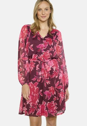 Robe florale avec un fond marron et de grandes fleurs roses, manches longues en tissu transparent, devant avec boutons et taille ceinturée.