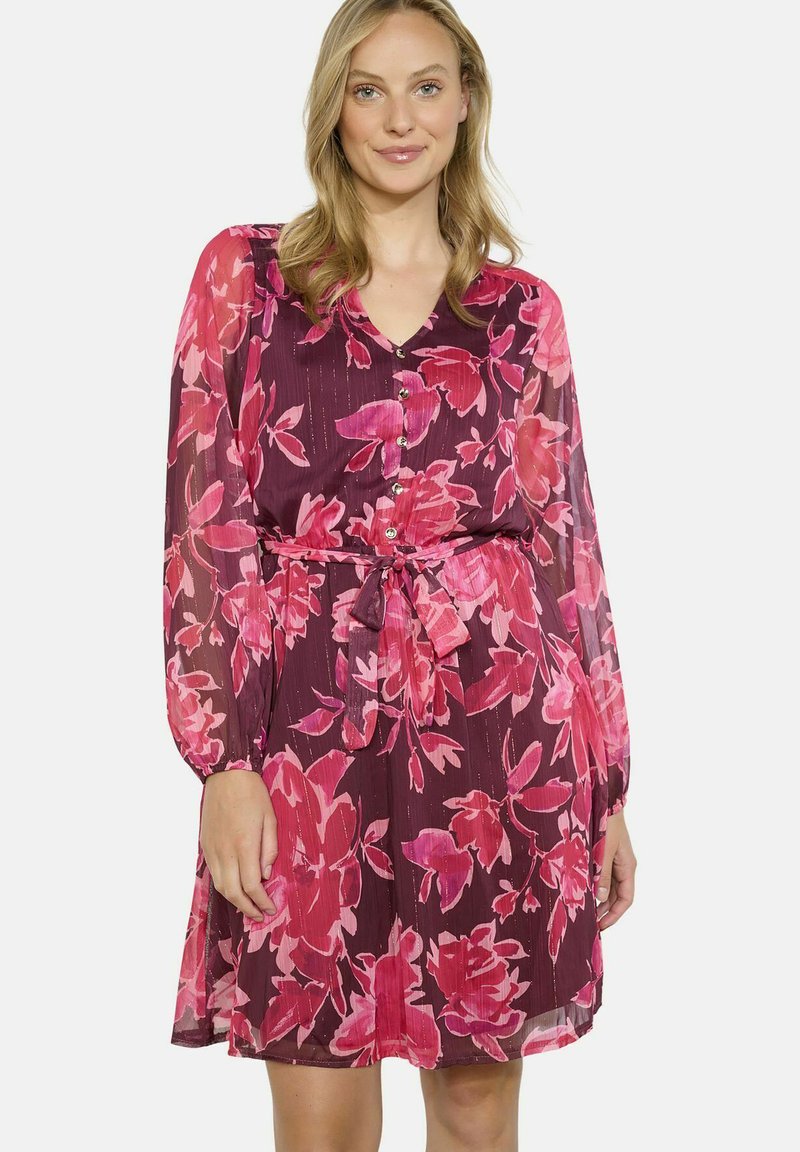 Vestido floral con un fondo marrón y grandes flores rosas, mangas largas y transparentes, un cierre frontal con botones y una cintura ceñida con cinturón.