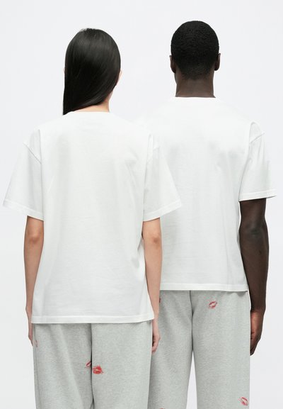 White cotton t-shirts with short sleeves, featuring a crew neck. Plain design, worn with gray sweatpants adorned with red lips pattern.