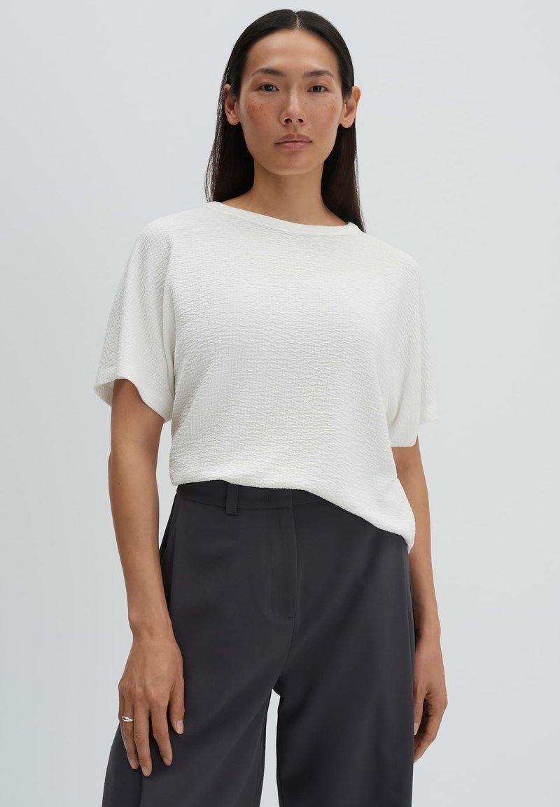 Femme aux cheveux longs et foncés portant un top blanc texturé à manches courtes et un pantalon noir taille haute, debout devant un fond clair uni.