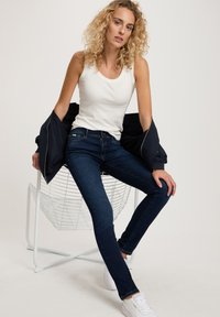 White tank top, dark blue jeans, and white sneakers. Sitting on a white wire chair, with a navy jacket draped over one arm.