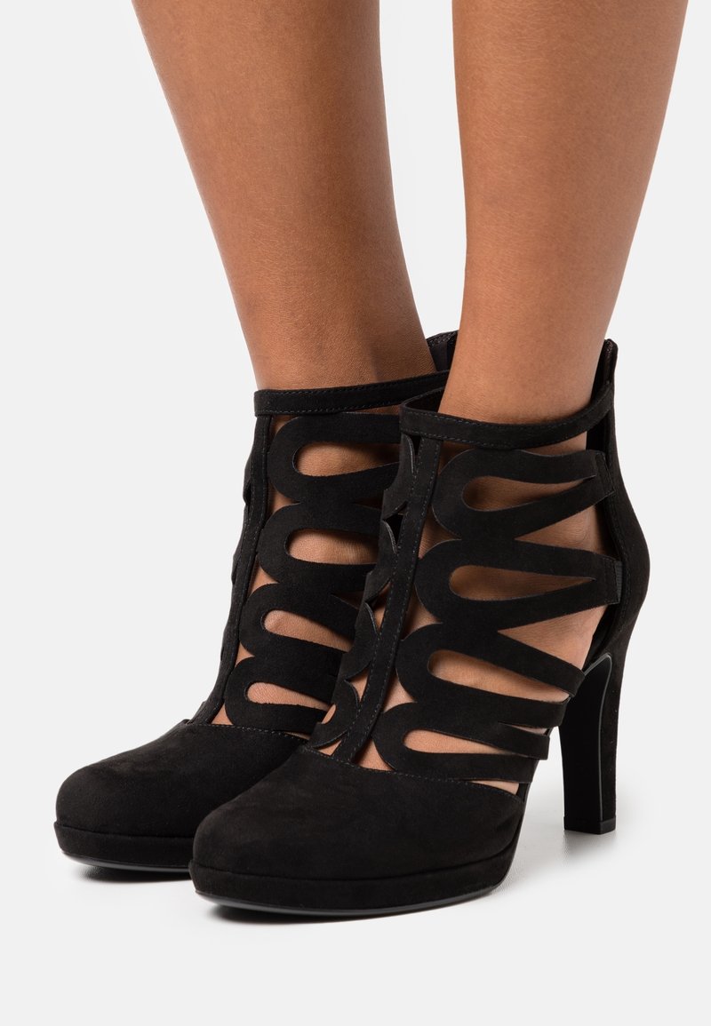 Chaussures à talons hauts en daim noir avec des brides découpées et des fermetures à la cheville, portées pieds nus sur un fond blanc.