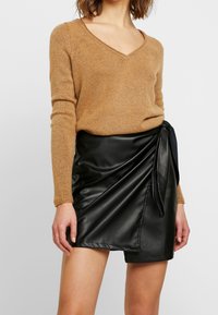 Pull marron texturé avec un col en V, associé à une jupe portefeuille en simili cuir noir dotée d'un nœud sur le côté et d'un design drapé.