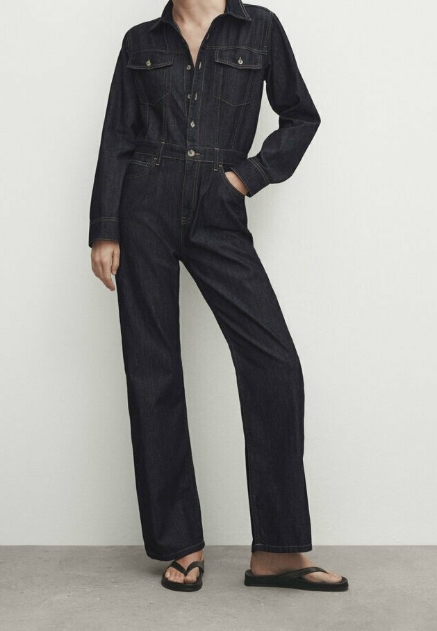 Kobieta w ciemnoniebieskim, długim kombinezonie z długimi rękawami, posiadającym kieszenie z przodu oraz zapinanym na guziki, stoi na tle w neutralnych kolorach. Ma na sobie czarne sandały.