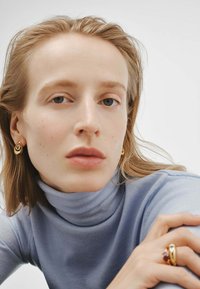 Mujer de piel clara y cabello rubio que lleva pendientes y anillo de oro, vestida con un suéter de cuello alto azul claro, mirando directamente al frente.