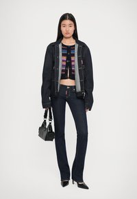 Chaqueta de mezclilla oscura sobre un cárdigan corto y colorido, combinada con jeans oscuros acampanados y tacones negros de punta. Se incluye un pequeño bolso negro.