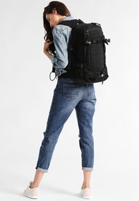 Musta reppu, jossa on säädettävät hihnat, useita etutaskuja ja bungee-nauhoja. Yhdistettynä vaaleaan denim-takkiin ja sinisiin farkkuihin.