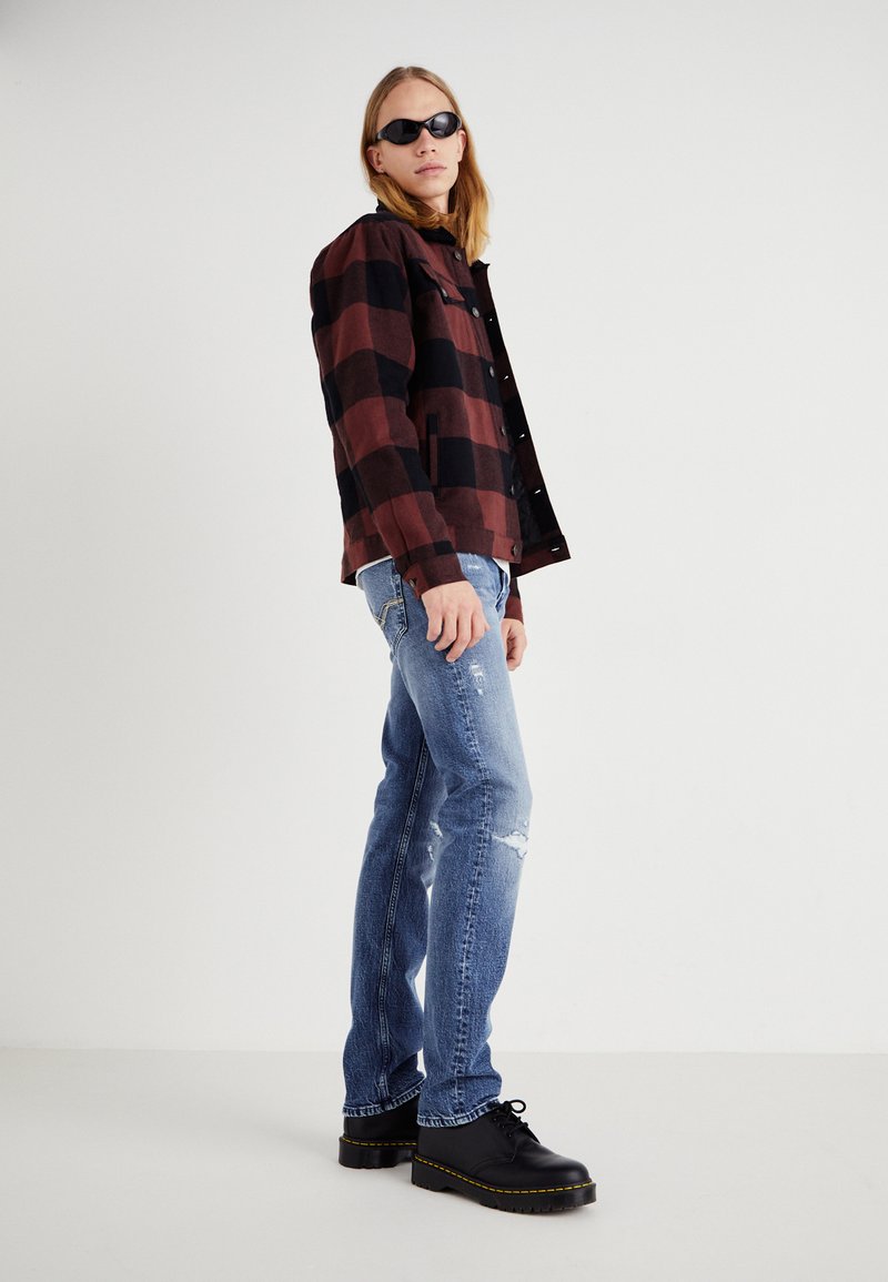 Zwart-rood geblokt shirtjack, blauwe versleten jeans en zwarte platform schoenen met gele stiksel, gedragen met zwarte zonnebril.