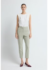 Vrouw staat tegen een witte achtergrond, gekleed in een mouwloze witte blouse, lichtgroene op maat gemaakte broek, gouden creolen en bordeauxrode lage hakken.