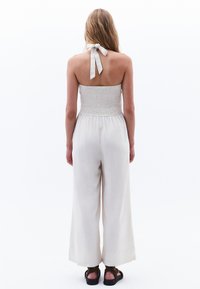 Lätt, krämfärgad jumpsuit med smockad byst, vida ben och halterneck-uppbindning. Mjuk textur med minimalistiska design detaljer.