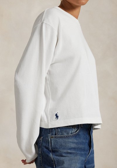Person iført en hvid langærmet top med et lille navy polo spillerlogo nær sømkanten og blå denimjeans mod en neutral baggrund.