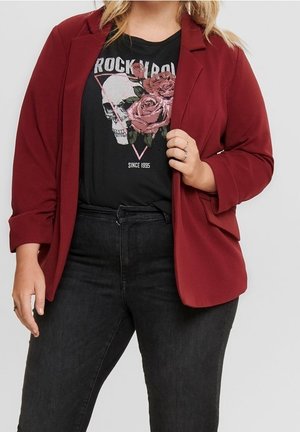 Kobieta nosząca czerwoną marynarkę, czarne jeansy z wysokim stanem oraz czarny t-shirt z grafiką czaszki, róż i napisem „Rock N Roll Since 1995”.