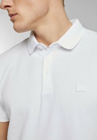 Vit polo t-shirt i strukturerad tyg, med krage, två-knapplåst och en liten präglad logotyp på vänster sida.