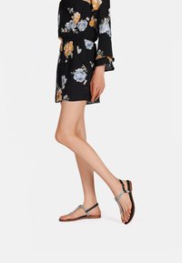 Vestito nero a fiori con maniche lunghe, caratterizzato da fiori blu e arancioni. Le calzature sono sandali neri con suola piatta, con un accento argento e una suola color tan.