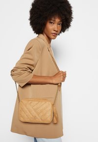 Beige quiltad crossbody-väska med en fransdetalj, tillverkad av ett strukturerat mockaliknande material och en guldkedjesträng.