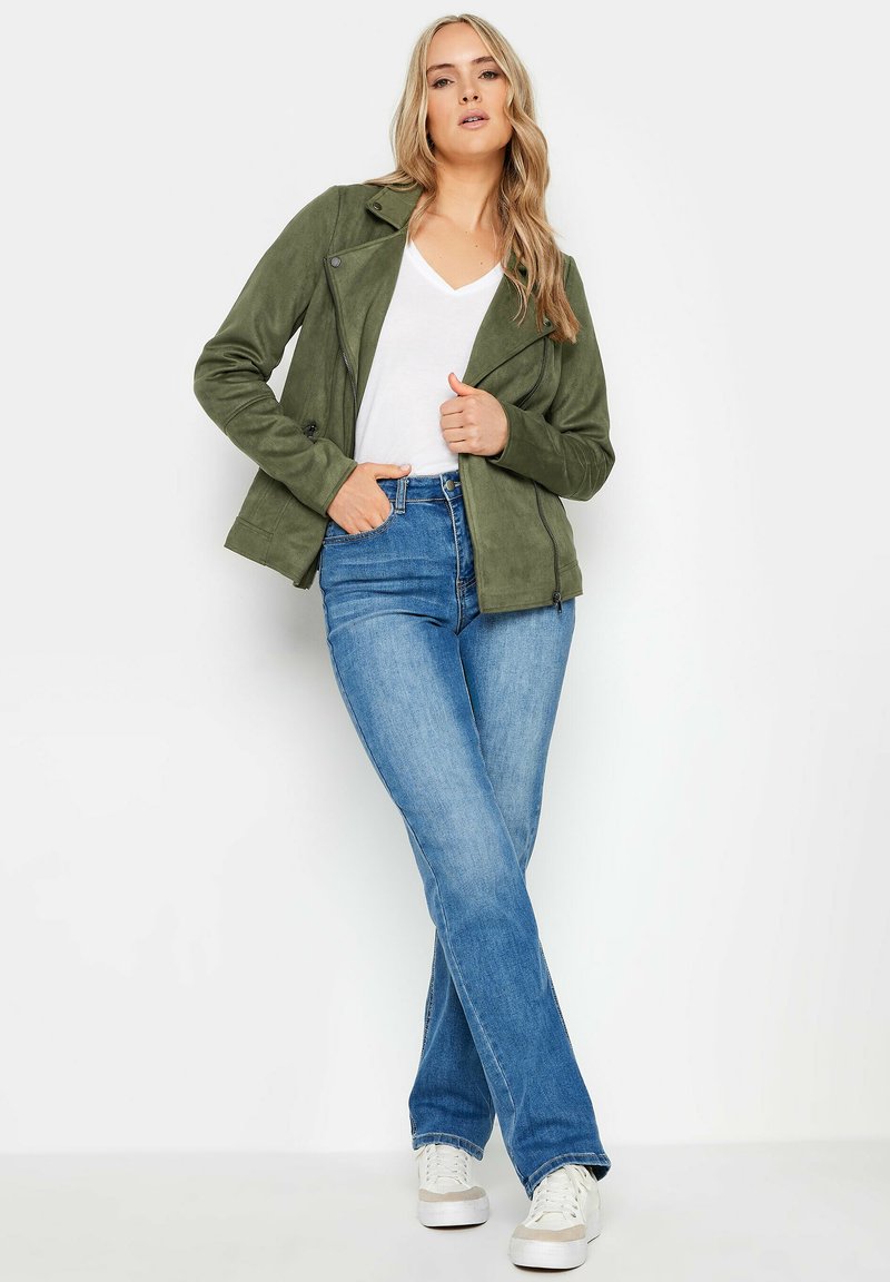 Olivgrön mockajacka med asymmetrisk dragkedja, figursydd design, i kombination med blå jeans och vita sneakers. Avslappnad, modern stil.