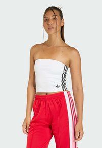 Biely bezramenný crop top s čiernymi bočnými pruhmi a malým logom, skombinovaný s červenými športovými nohavicami s bielymi bočnými pruhmi. Hladká textúra.