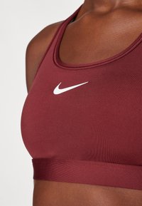 Bordo sportski grudnjak s bijelim Nike logom. Ima glatku tkaninu, okrugli izrez i elastični pojas na dnu. Čist, minimalistički dizajn.