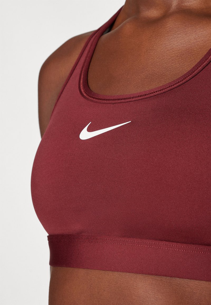 Sottoflessione sportiva color mogano con logo Nike bianco. Presenta un tessuto morbido, scollatura rotonda e un elastico nella parte inferiore. Design pulito e minimalista.