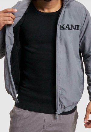 Homme portant une chemise noire et une veste zippée grise avec le texte "KANI", tenant la veste ouverte pour montrer la doublure intérieure.