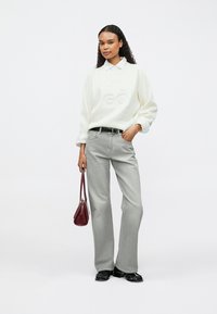 Bela majica z izbočenim logotipom, sive razširjene jeans hlače, črni čevlji in majhna bordo torbica. Model stoji z eno roko na boku.