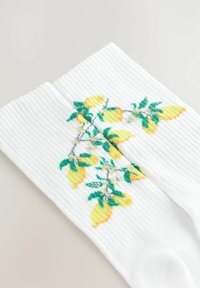 Hvide ribbede sokker med et farverigt design af citroner og grønne blade. Mønsteret inkluderer gule citroner og små hvide blomster.