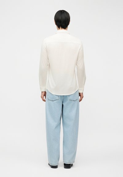Άτομο που στέκεται πλάτη, φοράει λευκό πουκάμισο με μακριά μανίκια και ανοιχτό μπλε τζιν παντελόνι με φαρδιά μπατζάκια, καθώς και μαύρα παπούτσια, σε απλό φόντο.