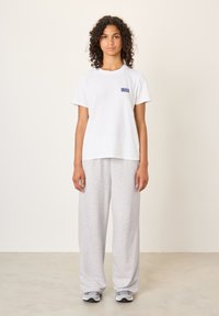 Balta kokvilnas T-krekls ar zilu "American Vintage" logotipu, papildināts ar gaiši pelēkām, vaļīgām sporta biksēm un pelēkām sporta kurpēm.