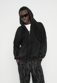 Μαύρο φούτερ με φερμουάρ και κουκούλα, που διαθέτει φθαρμένες λεπτομέρειες και δύο μπροστινές τσέπες. Συνδυάζεται με σκούρο παντελόνι με αφηρημένο γκρι μοτίβο.