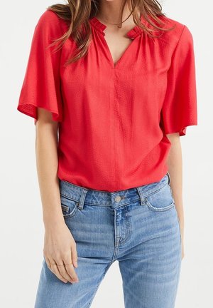 Kvinde iført en knaldrød kortærmet bluse med flagrende ærmer, kombineret med lyseblå denimjeans, hånd hvilende nær lommen.