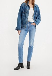 Chaqueta de mezclilla en azul claro, de corte relajado, con cierre de botones y dos bolsillos en el pecho. Combinada con vaqueros azul claro y botas de tobillo negras de tacón.