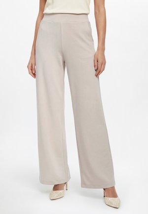 Kobieta w beżowych spodniach o szerokich nogawkach, kremowej bluzce bez rękawów i beżowych szpiczastych szpilkach z ozdobami, stojąca na białym tle.