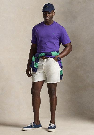 Mand iført en lilla t-shirt, hvide shorts, navy kasket, navy sneakers og en grøn-og-navy sweater bundet om taljen, står foran en neutral baggrund.