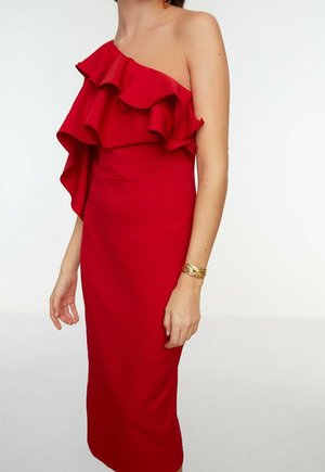Kobieta ubrana w czerwoną sukienkę midi na jedno ramię z falbankami, uzupełnioną złotą bransoletką na nadgarstku.